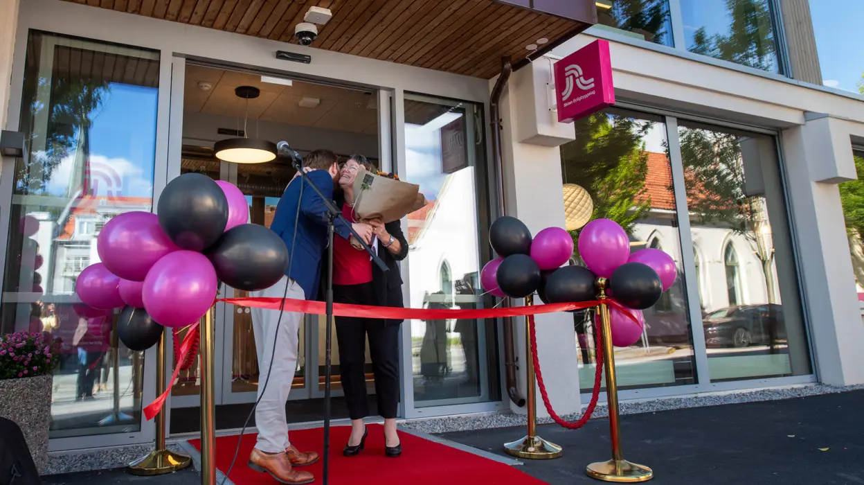 En mann klemmer en dame og gir blomster. Utenfor ett bygg med ballonger, rød løper og snor, klar for å bli klippet.