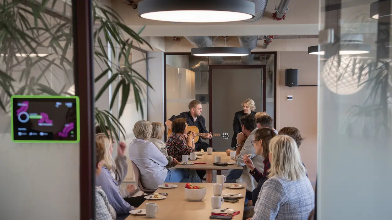 En mann med guitar og en dame synger og spiller for en gruppe mennesker i et konferanserom