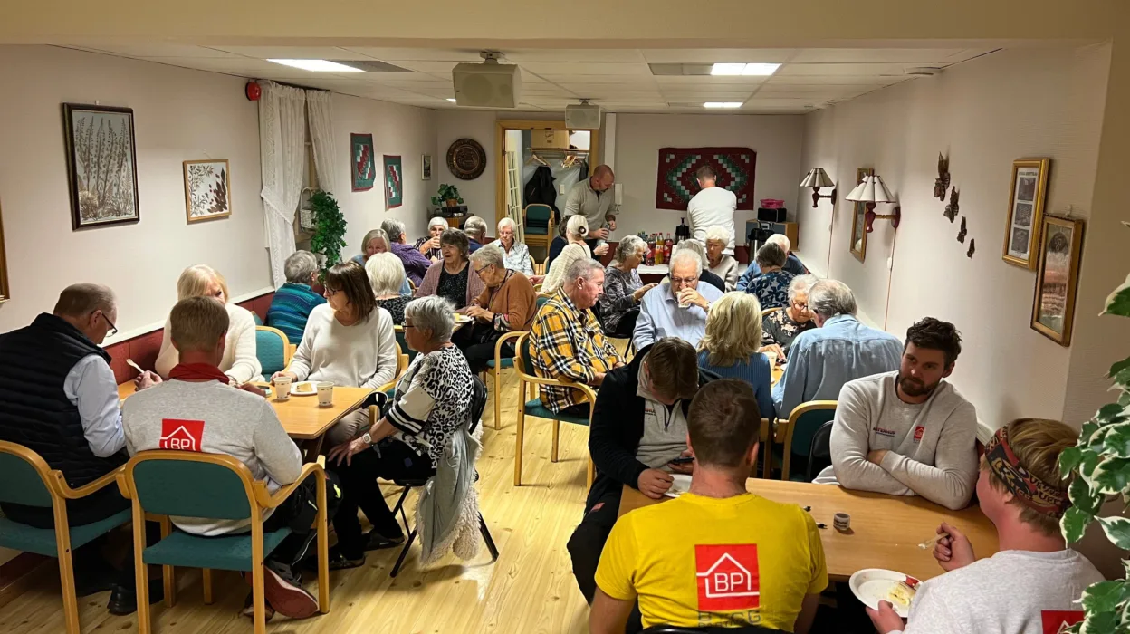 Stor gruppe med folk fordelt på mange bord. De spiser kake.