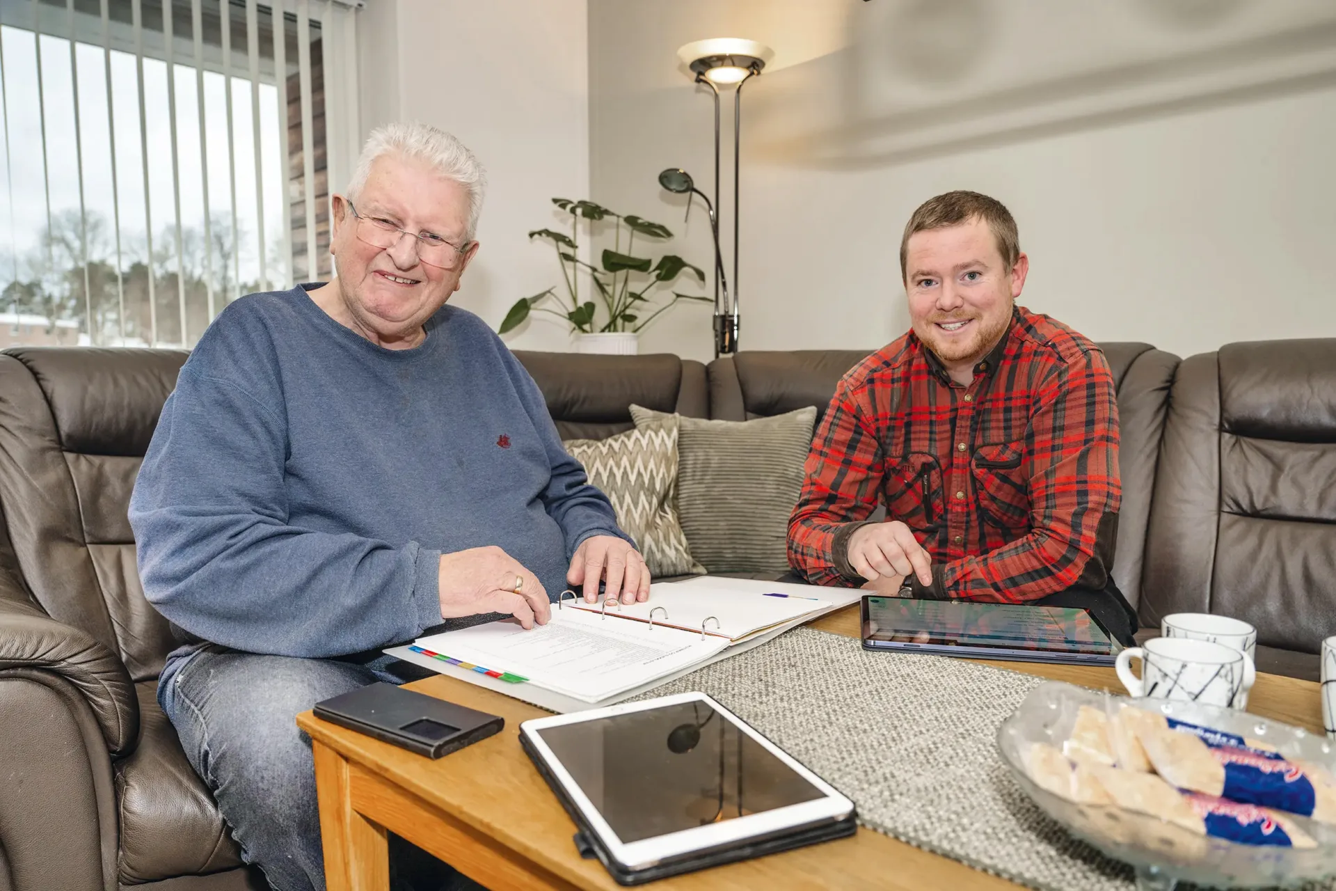 Em eldre og en yngre mann sittende i sofa ved bord med permer og nettbrett. lig i Arendal Boligbyggelag Talleiv Midtgarden